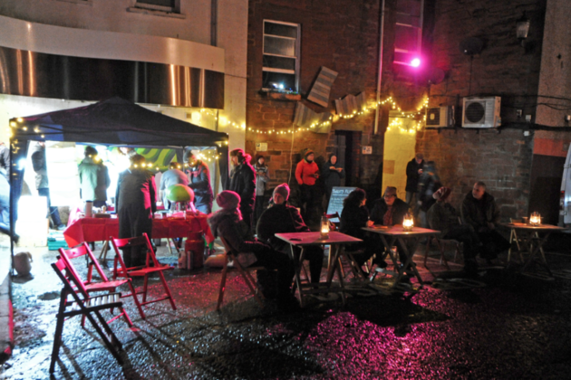 A number of fold out tables and chairs sit in the street in the dark. People sat in the chairs are chatting. A string of fairy lights and purple spotlights light the scene. 